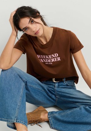 Ung kvinde med mørkt hår, der sidder med benene krydset, iført en brun "Weekend Wanderer" t-shirt, blå jeans og lyse snøresko mod en ensfarvet baggrund.