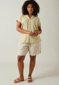 Blusa de botones de manga corta en amarillo con un patrón geométrico blanco, combinada con pantalones cortos beige claros que tienen dos bolsillos frontales. Sandalias marrones.