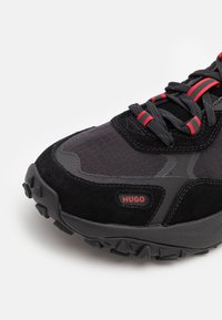 Chaussure de sport noire associant des matériaux lisses et texturés, avec des accents rouges sur les lacets et une semelle extérieure robuste dotée de motifs d'adhérence.