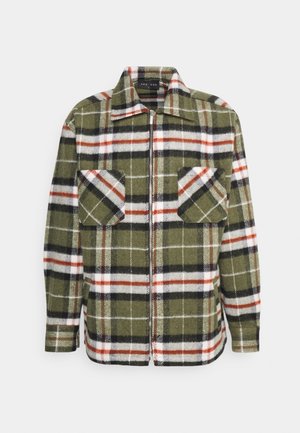 Groene geruite flanellen shirt met rode en witte accenten, over een donkere hoodie gedragen. Zwarte broeken en zwarte sportschoenen maken de outfit compleet.