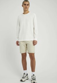 Chemise blanche à manches longues, shorts beiges retroussés, et baskets épaisses avec des accents multicolores, se tenant contre un fond neutre.