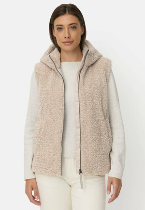 Kvinde iført en beige, fluffy, lynlåsfat, ærmeløs vest med hætte over en lys, langærmet top og lyse bukser.