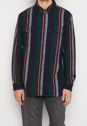 Homme portant une chemise à manches longues à rayures verticales bleu marine, vert, bordeaux et gris, associée à un jean gris foncé.