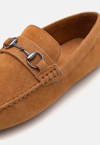 Brune ruskindsloafer med en dekorativ metalaccent, tekstureret overflade og syede detaljer langs kanterne. Blødt indvendigt foer er synligt.