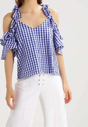 Blusa a quadri blu e bianchi con maniche a sbuffo scollate e scollo a V, realizzata in tessuto leggero. Abbinata a pantaloni bianchi.