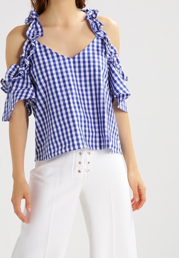 Blå och vit rutigt blus med avskurna volangärmar och V-ringning, tillverkad av lättviktsmaterial. Matchad med vita byxor.