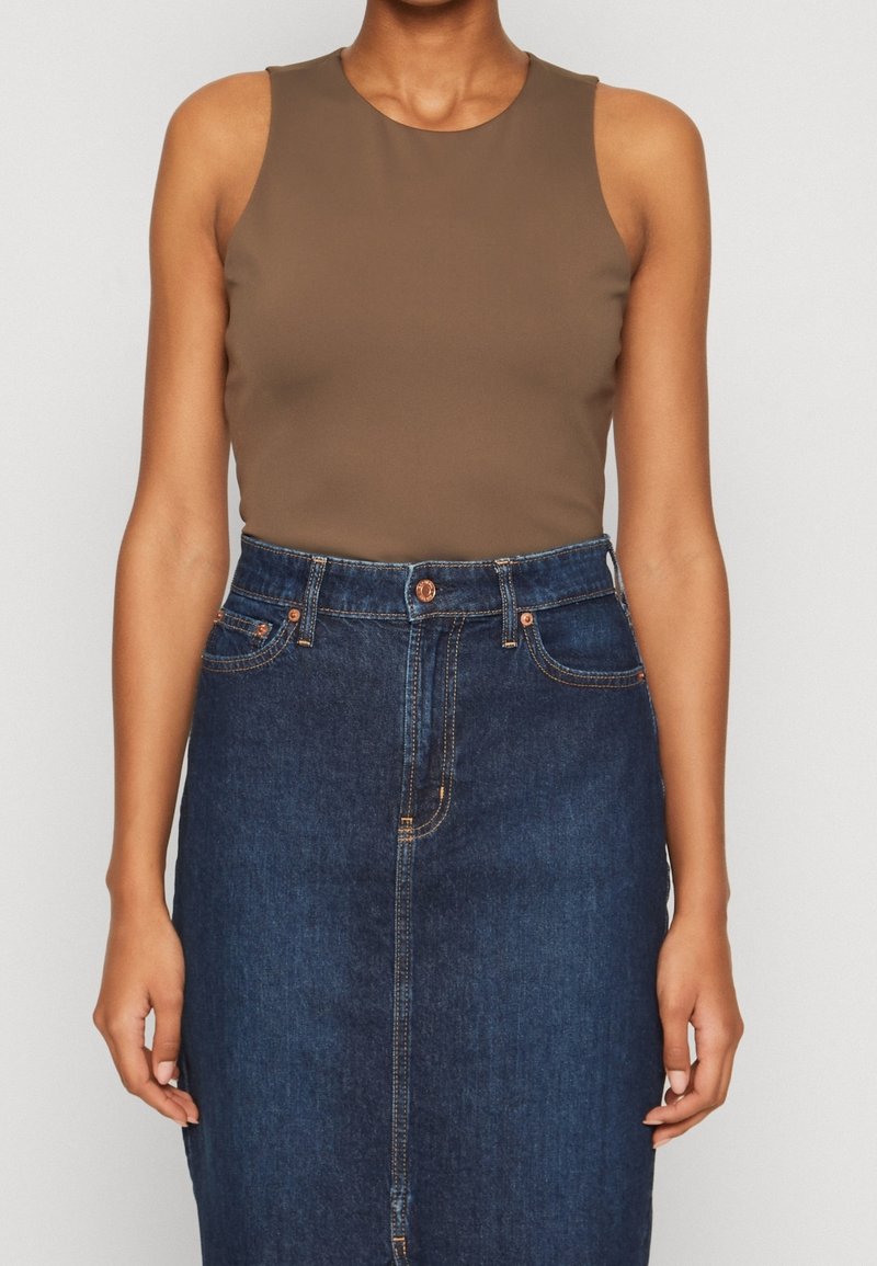 Femme portant un haut marron sans manches rentré dans une jupe en denim bleu foncé taille haute avec des boutons et des poches à l'avant.
