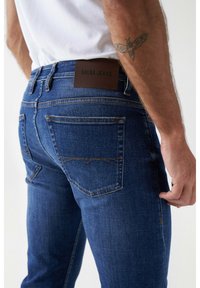 Tummansiniset farkut, jotka on valmistettu denimmateriaalisesta, tiukasti istuvasta leikkauksesta. Niissä on nahkainen merkki, jossa lukee "SALSA JEANS", sekä yksityiskohtainen stitchaus takataskuissa.