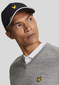 Cappello nero con logo dell'aquila gialla, dettagli bianchi e un maglione grigio a scollo a V con logo ricamato. Tessuto dalla texture liscia.