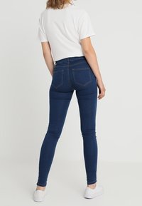 Los jeans skinny de mezclilla azul presentan un diseño de cintura alta, bolsillos traseros y una textura suave, combinados con una camiseta blanca y zapatillas ligeras.
