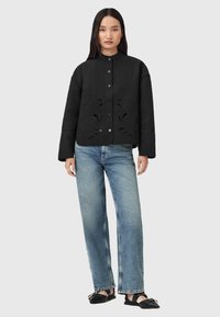 Donna in piedi che indossa un giubbino nero imbottito con motivi a intaglio, jeans azzurri a gamba dritta e sandali neri flat con fibbie.