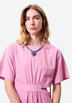 Jeune femme aux cheveux blonds mi-longs portant une robe rose et un collier en perles bleues en forme de cœur, faisant face à l'avant avec une expression neutre.