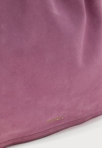 Τσαντάκι ώμου από μωβ σουέντ με εμφανές χρυσό λογότυπο "Coccinelle" κοντά στην κάτω άκρη, σε λευκό φόντο.