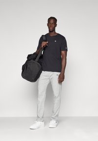 Schwarzes T-Shirt mit kurzen Ärmeln und Logo, graue Sweatpants, weiße Sneaker und eine schwarze Sporttasche, die vor einem hellgrauen Hintergrund steht.