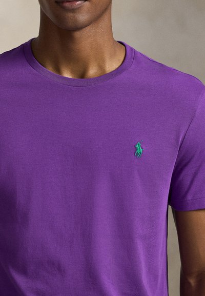 Μωβ βαμβακερό μπλουζάκι με στρογγυλή λαιμόκοψη και κοντά μανίκια, με ένα μικρό πράσινο κεντημένο λογότυπο στο αριστερό στήθος. Ομαλή υφή.