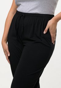 Schwarze Stoffhose mit elastischem Bund und Zugband, aus weichem Material gefertigt. Verfügt über seitliche Taschen und einen geraden Schnitt. Glatte Textur.