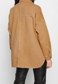 Giacca camicia in cotone marrone con vestibilità rilassata, maniche lunghe, polsini con bottoni e orlo curvo, abbinata a pantaloni in pelle nera.