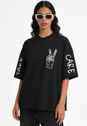 Donna con occhiali da sole rettangolari bianchi e una t-shirt nera oversize con un simbolo di pace e le parole "Faith," "Love," "Peace," e "Joy Hope."
