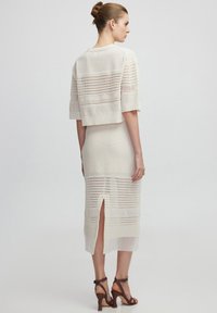 Strick-Zweiteiler in Creme, bestehend aus einem kurzhalsigen Top mit horizontalem Streifenmuster und einem Midi-Rock mit Schlitz an der Seite. Getragen mit braunen High Heels.