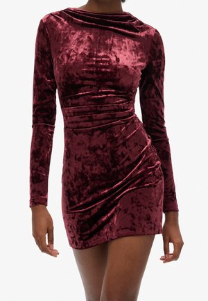 Woman wearing a long-sleeve, ruched, deep red velvet mini dress, standing against a plain white background.