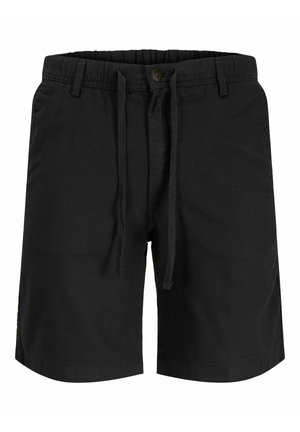 Dospievajúci chlapec stojaci, oblečený v béžovej košeli s dlhým rukávom, čiernych kraťasoch, bielych ponožkách po lýtka a bielych teniskách na jednoduchom sivom pozadí.