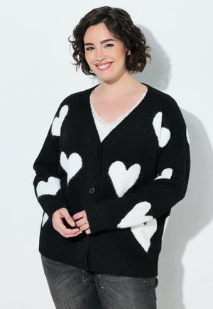 Kvinde med kort krøllet hår smiler iført en sort cardigan med store hvide hjerter over en hvid top og grå bukser.