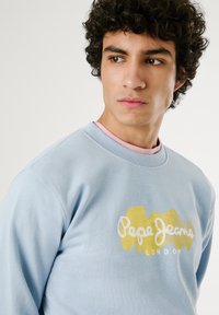 Lichtblauwe sweatshirt met ronde halslijn, voorzien van een wit en goud logo ontwerp. Zachte katoen material met een ontspannen pasvorm.