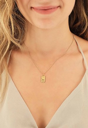 Collier en pendentif en or avec un charme rectangulaire, présentant un motif texturé et une chaîne fine, porté sur peau nue.