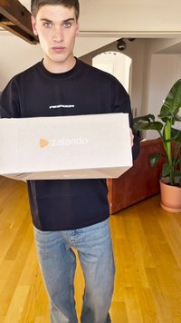 Kartonowe pudełko Zalando w jasnobrązowym kolorze z pomarańczowym logo. Osoba ubrana w czarną bluzę z długim rękawem i jasnoniebieskie dżinsy, stojąca na drewnianej podłodze.