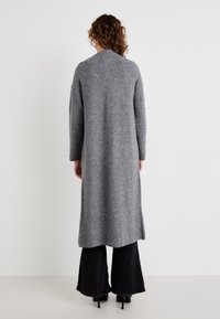 Lang grå strikket cardigan med ribbestruktur, åpen front og sidespalter, kombinert med svarte vide bukser. Ingen synlige lukkinger.