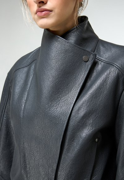 Vrouw draagt een zwart lederen jas met een hoge kraag en textuur, voorzien van een verborgen drukknoopsluiting.