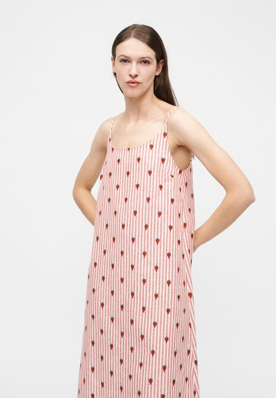 Γυναίκα με μακριά καστανά μαλλιά φοράει αμάνικο φόρεμα με κάθετες κόκκινες και άσπρες ρίγες και μικρό σχέδιο φράουλας, στέκεται με τα χέρια πίσω.