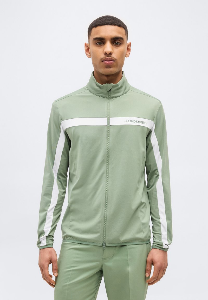 Άνδρας που φοράει ανοιχτοπράσινο μπουφάν με φερμουάρ και λευκές ρίγες στα μανίκια και το στήθος, στέκεται μπροστά σε απλό φόντο.