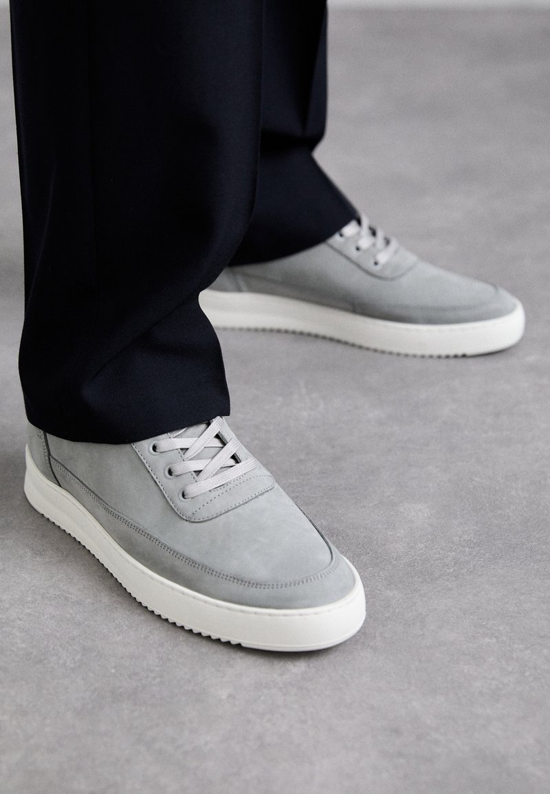 Zapatillas de ante gris con suelas de goma blanca, que cuentan con un diseño de cordones, punta redondeada y detalles de costura sutiles, llevadas con pantalones negros.