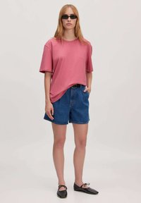 Roze oversized katoenen t-shirt, gecombineerd met blauwe denim shorts. Zwarte platte schoenen met een vetersluiting. Model draagt een paar donkere zonnebrillen.