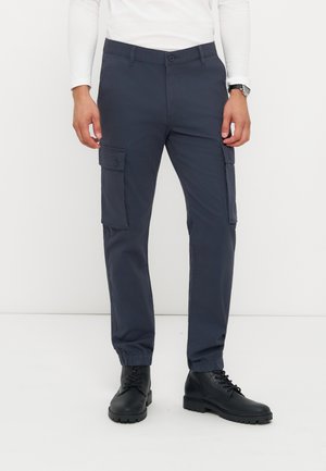 Homme portant un pantalon cargo bleu foncé avec des poignets élastiques, des bottes noires à lacets, une chemise blanche à manches longues et une montre-bracelet au poignet gauche.