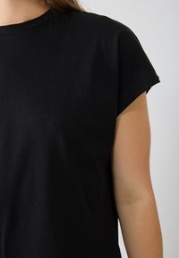 T-shirt en coton noir avec col rond et manches courtes. Le tissu semble lisse avec un design minimaliste et sans motifs ni accents visibles.