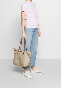 Bolso tote beige con asas de cuero marrón, con una textura suave y herrajes dorados. Combinado con una camiseta de rayas rosa y blanca y jeans azules.