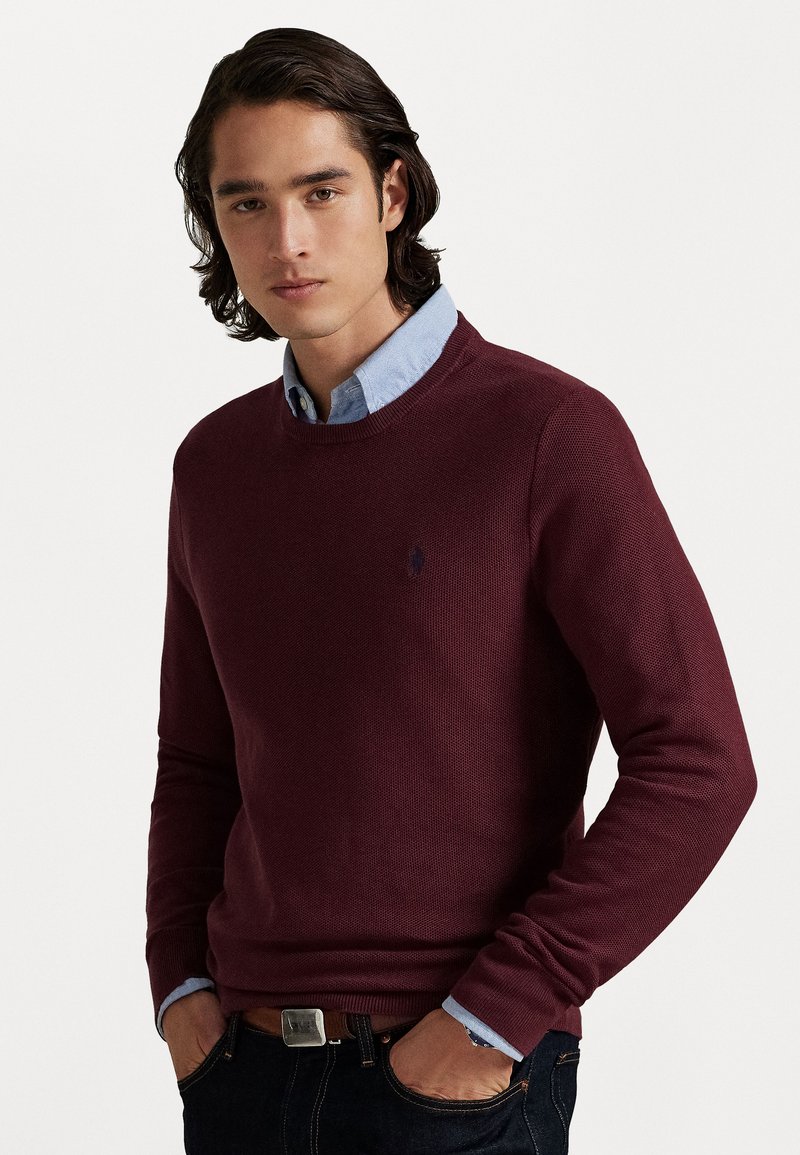 Joven con cabello oscuro de longitud media, vistiendo un suéter burdeos sobre una camisa de cuello azul claro, con las manos en los bolsillos de unos jeans oscuros.