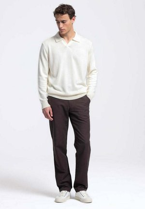 Man draagt een witte lange mouwen coltrui, donkere broek en witte sneakers, staat met één hand in de zak en kijkt naar beneden.