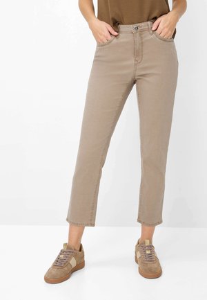 Person iført beige kortere bukser og beige sneakers med hænderne i lommerne mod hvid baggrund.