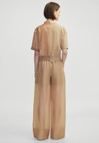 Hellbrauner Jumpsuit mit kurzen Ärmeln, elastischem Bund und weitem Bein. Transparente Textur mit glatter Oberfläche.