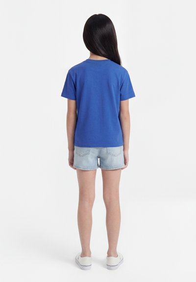 Laps seisab seljaga vaataja poole, seljas ühevärviline sinine lühikeste varrukatega särk, heledad teksase lühikesed püksid ja valged libisevad jalanõud valgel taustal.