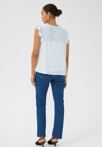 Blouse sans manches bleu clair avec des épaules froncées, associée à un jean en denim bleu foncé et des mules à talons noirs, vue de dos.