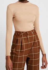 Top lungo a maniche lunghe a coste beige con collo alto, abbinato a pantaloni a quadri marroni con accenti bianchi e dettagli plissettati.