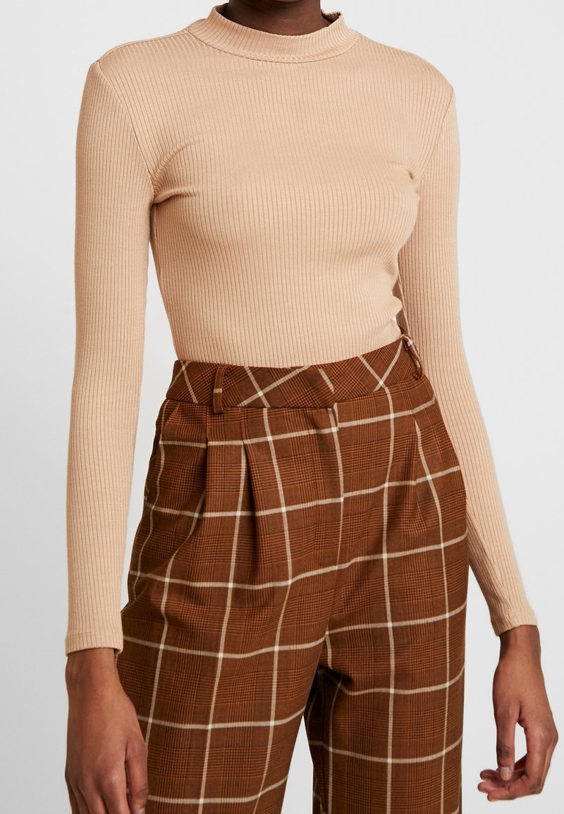 Top lungo a maniche lunghe a coste beige con collo alto, abbinato a pantaloni a quadri marroni con accenti bianchi e dettagli plissettati.