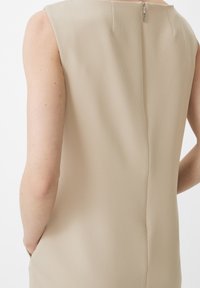 Top sin mangas beige con un tejido texturizado, que cuenta con una cremallera en la parte trasera, bolsillos laterales y una silueta ajustada.