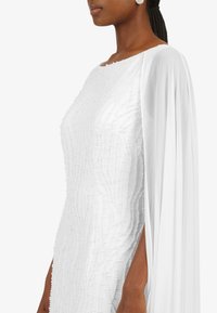 White gown with textured beading, a smooth draped cape, and side slit. Features a boat neckline and is fitted at the waist.