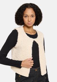 Cremfarbener, flauschiger Weste mit rundem Ausschnitt, ohne Verschlüsse und offener Vorderseite. Über einem schwarzen Langarmshirt gestylt, das eine weiche Textur aufweist.