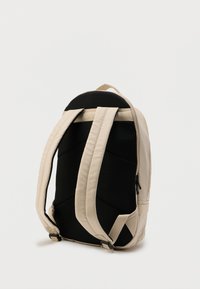 Μπεζ σακίδιο πλάτης με επενδυμένες λουρίδες ώμου και μαύρο μαξιλαρωτό πίσω πάνελ, όρθιο σε λευκή απλή επιφάνεια.